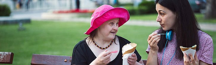 Zwei Frauen sitzen auf einer Parkbank und essen Eis, eine hat eine sichtbare Behinderung